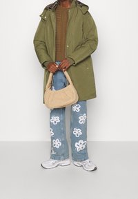 Persona con parka verde olivo, suéter de punto marrón, jeans azules con estampados de flores blancas, zapatillas blancas y sosteniendo un bolso de pelo beige.