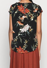 Haut noir à motifs floraux avec des manches courtes, fermeture éclair au dos et ourlet arrondi. Le motif comprend des fleurs rouges, blanches et vertes.