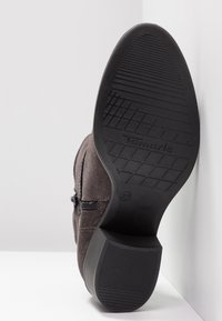 Semelle droite d'une botte en suède marron foncé avec une semelle extérieure en caoutchouc noir texturé et une fermeture éclair latérale, inclinée contre un mur blanc en angle.