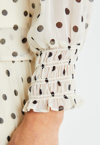 Tessuto cremoso trasparente con pois neri; presenta polsini arricciati con dettagli a ruches. Leggero e testurizzato, adatto per look a strati.