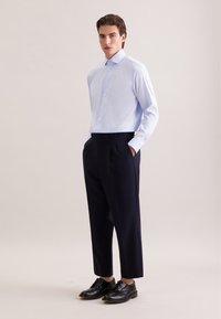 Lichtblauw overhemd met lange mouwen en knoopsluiting, gecombineerd met een high-waist zwarte broek en zwarte leren schoenen. Neutrale achtergrond.