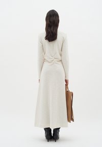 Lång crème-färgad klänning i en texturerad tyg, med en rynkad detalj på baksidan, tillsammans med en brun tote-väska och svarta klackstövlar.