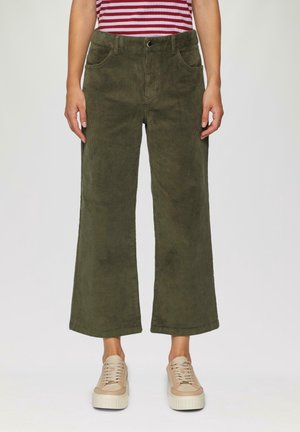 Person trägt olivgrüne Cordhose mit weitem Bein, beige Plateausneaker und ein rot-weiß gestreiftes Oberteil, steht vor einem schlichten Hintergrund.