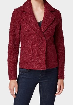 Kvinde iført en tekstureret dybrød skræddersyet jakke med revers over en sort top, kombineret med mørkeblå jeans, stående med armene afslappede.