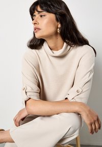 Top beis de cuello simulado hecho de tela texturizada, con mangas enrolladas y botones. El modelo se sienta con pantalones de un beige claro y una silla de madera.
