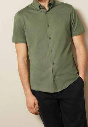 Homme portant une chemise boutonnée vert olive à manches courtes et un pantalon noir, avec une main dans la poche, sur un fond beige.