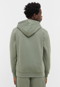 Mężczyzna odwrócony plecami, ubrany w jasną oliwkową bluzę z kapturem oraz dopasowane spodnie na tle czystej białej ściany.