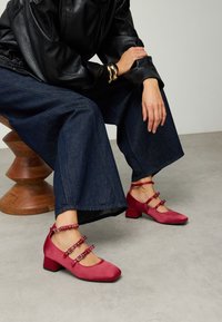 Chaussures rouges à bout carré, à talon bas, dotées de trois brides à la cheville ornées d'accents métalliques ; portées avec un pantalon en denim à jambes larges et un haut en cuir noir.