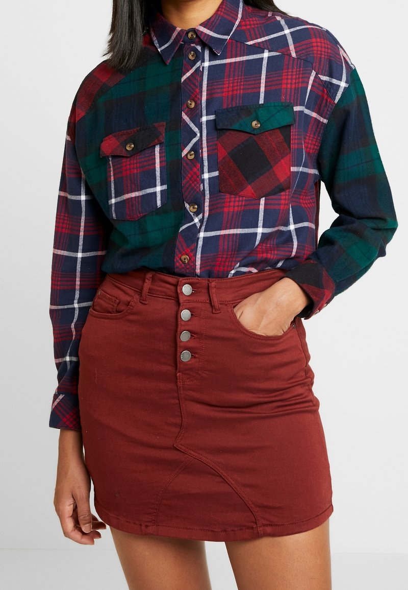 Chemise en flanelle à carreaux verts, rouges et bleu marine avec boutons et deux poches poitrine, associée à une jupe en denim bordeaux à cinq boutons.