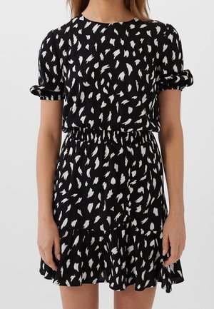 Vrouw draagt een zwart jurkje met korte mouwen, voorzien van een wit abstract penseelstreekpatroon en een elastische taille, staand tegen een eenvoudige achtergrond.