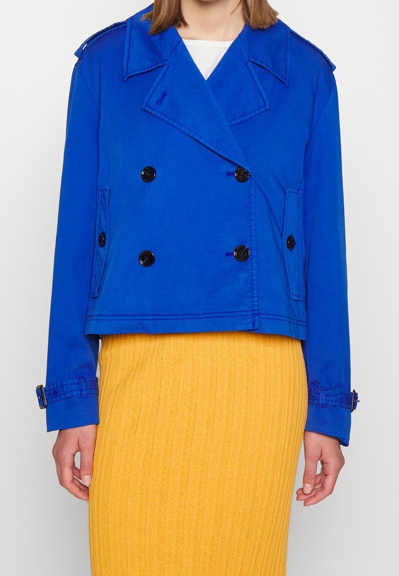 Personne portant une veste bleu vif à double boutonnage avec des boutons noirs sur une jupe jaune côtelée, devant un fond uni.