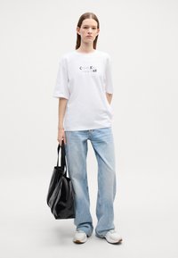 Witte katoenen T-shirt met zwarte opdruk "Calvin Klein Since 1968". Losse pasvorm, korte mouwen gecombineerd met lichtblauwe denim jeans en een zwarte tote bag.