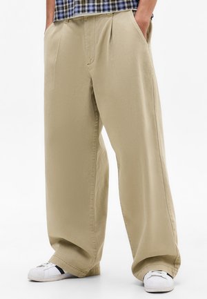 Pantaloni beige a gamba larga con pieghe, realizzati in tessuto morbido. Presentano tasche laterali e una vestibilità rilassata, abbinati a sneakers bianche.