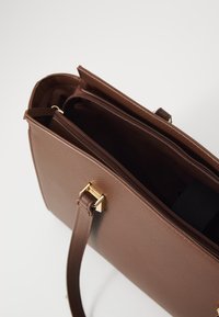 Bolso de hombro de cuero sintético marrón con cierre superior de cremallera, que presenta herrajes en tono dorado y un bolsillo interior. Textura suave, diseño estructurado.