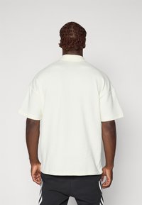 Polo majica s kratkimi rokavi v svetlo rumeni barvi z teksturiranim vzorcem, opremljena s kragnjo in ohlapnim krojem, v kombinaciji s črnimi trenirkami z črtami.