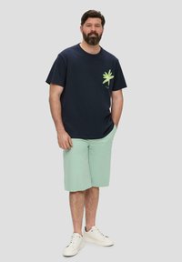 T-shirt en coton navy avec un graphique de feuille verte, associé à un short vert menthe et des baskets blanches. Un fond uni améliore la visibilité.