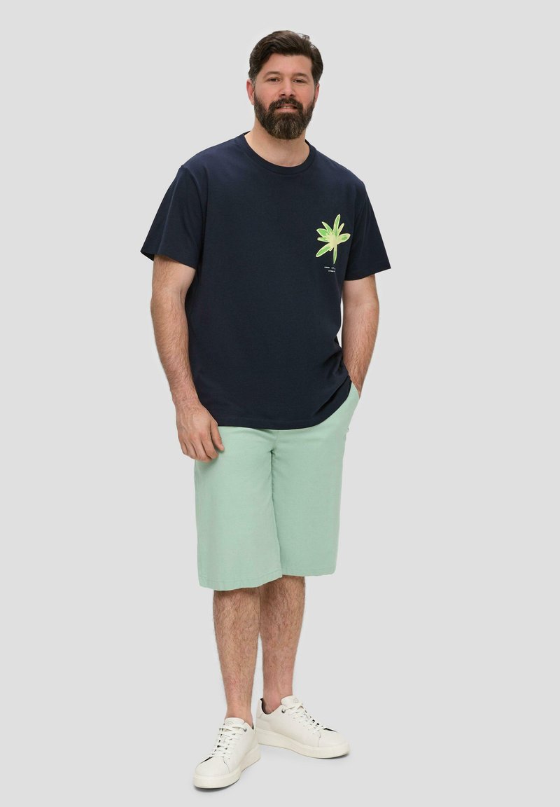 T-shirt en coton navy avec un graphique de feuille verte, associé à un short vert menthe et des baskets blanches. Un fond uni améliore la visibilité.