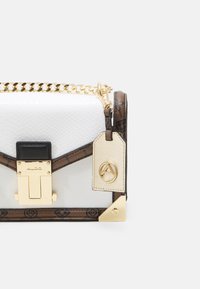 Bílá kabelka z umělé hadí kůže s hnědými detaily, zlatým kováním a řetízkovým popruhem. Má přední zapínání a ozdobnou visačku.