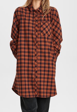 Vrouw draagt een geblokte jurk met lange mouwen in bruin en zwart, met knopen aan de voorkant en een borstzak, gecombineerd met een zwarte broek.