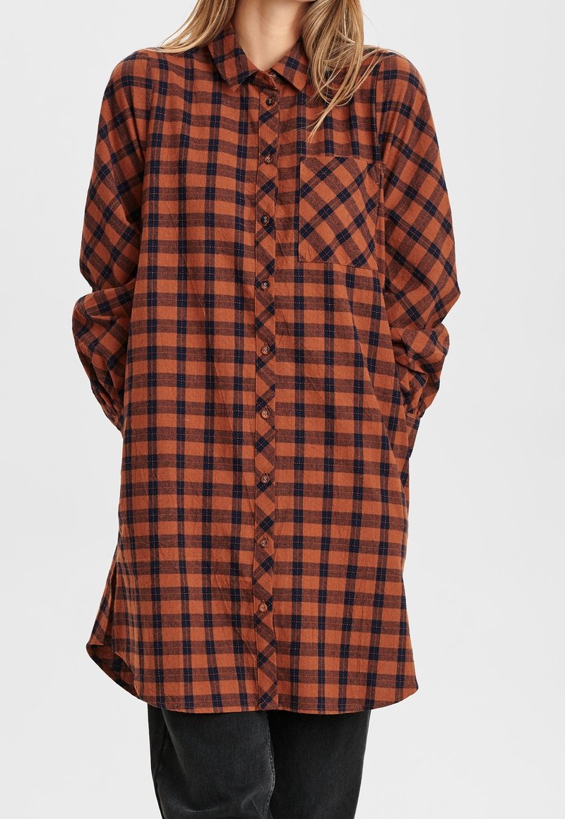 Femme portant une robe chemise à manches longues à carreaux marron et noir avec des boutons à l'avant et une poche poitrine, associée à un pantalon noir.