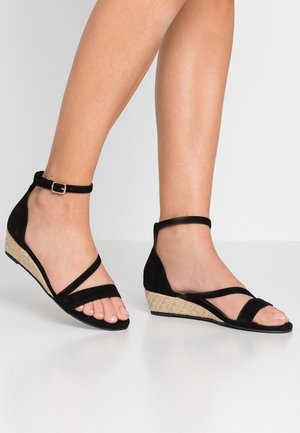 Fødder iført sorte kilehæls-sandaler med ankelremme og flettede beige hæle på en lysegrå gulv og baggrund.