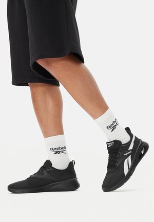 Czarne buty sportowe z siateczkową tkaniną, zaokrąglonym noskiem i białymi akcentami. Noszone z białymi skarpetkami z czarnym logo Reebok i czarnymi spodenkami.