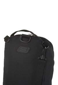 Bolsa de tela negra con cierre de cremallera, asa superior y correa ajustable. Presenta un logo moldeado y acentos de hardware metálico. Superficie texturizada.