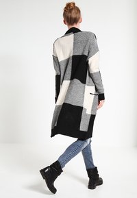 Cardigan bianco e nero con un design a patchwork geometrico, caratterizzato da una vestibilità lunga e rilassata e polsini a costine. Adatto per essere indossato a strati.