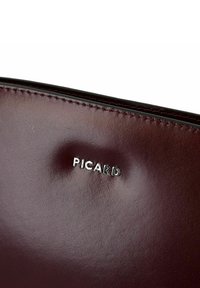 Bolso de cuero burdeos con textura suave, costuras en contraste y logotipo plateado en relieve. Diseño compacto y minimalista.