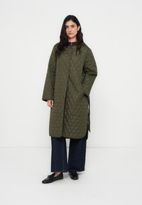 Πράσινο αμπιγιέ παλτό με καπιτονέ υφή σε μήκος μέχρι το γόνατο, άνετη εφαρμογή, με φερμουάρ και κούμπωμα με κουμπιά. Μακριά μανίκια και δύο πλαϊνές τσέπες.