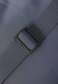 Tessuto grigio con una superficie liscia, caratterizzato da una fibbia scura e una robusta cinghia in tessuto. La vista ravvicinata mette in evidenza il materiale e i dettagli della cinghia.