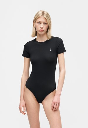 Blondínka v priliehavom čiernom body s krátkym rukávom a malým bielym logom na hrudi, stojaca pred jednoduchým svetlošedým pozadím.