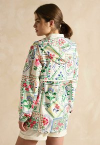 Jachetă ușoară cu modele florale, având o bază albă, cu accente florale colorate, glugă și manșete elasticizate.