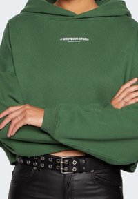 Persona che indossa un hoodie verde con la scritta bianca "WRSTBHVR STUDIO" e pantaloni di pelle nera con cintura nera a borchie incrocia le braccia in vita.