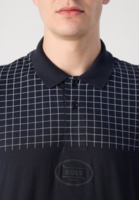 Čierne polo tričko s bielym mriežkovým vzorom a čiernou nášivkou loga. Má klasický golier a zapínanie na tri gombíky. Štruktúrovaná tkanina.