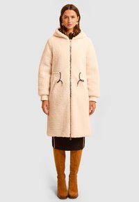 Beżowy płaszcz z polaru z miękkim kapturem, zamkiem błyskawicznym i dwoma przednimi kieszeniami. Posiada czarne troczki. Długie rękawy z ściągaczami.