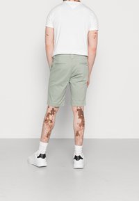 Shorts verts clairs avec une coupe plate et des poches arrière, associés à un t-shirt blanc. Le modèle porte des baskets blanches avec un accent noir.
