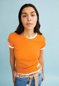 Oranje geribde t-shirt met witte accenten op de mouwen; gecombineerd met lichtblauwe jeans en een beige gestreepte omslag om de taille.