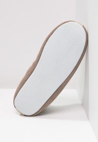 Brown slipper with a soft white sole, resting against a white corner wall on a flat surface.