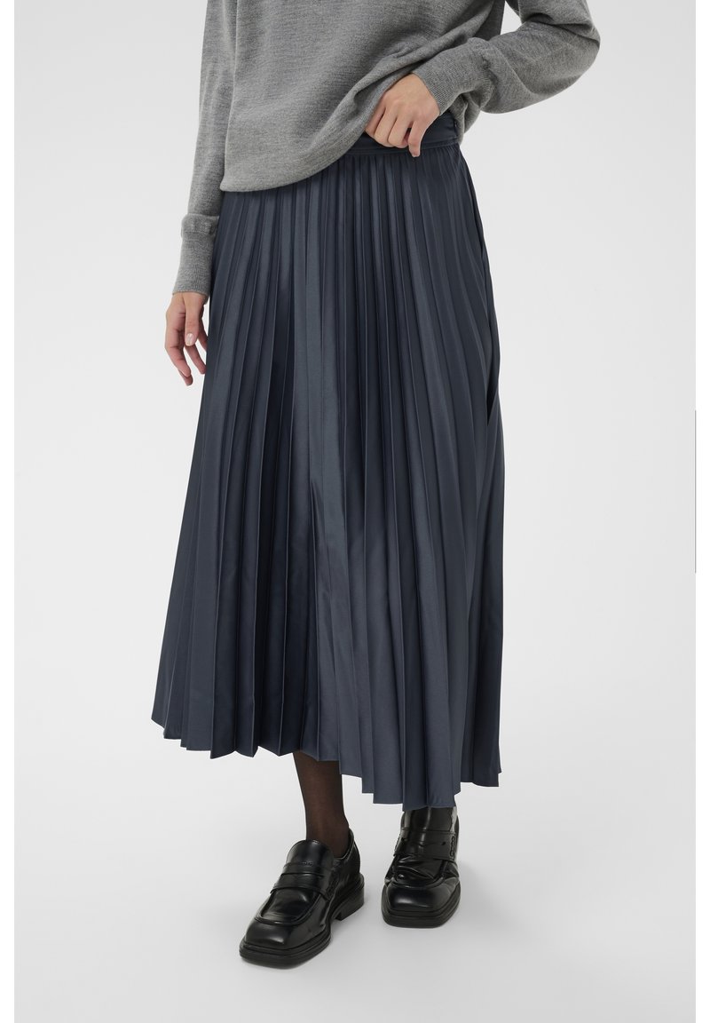 Gonna a pieghe blu navy realizzata in tessuto lucido, con vita alta e lunghezza fino a metà polpaccio. Abbinata a un maglione grigio e scarpe nere.