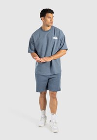 Camiseta de algodón azul de manga corta con cuello redondo, combinada con pantalones cortos a juego. El modelo lleva zapatillas blancas y calcetines de caña corta con un patrón.