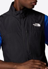Black sleeveless vest with high collar, front zip, and a small white logo on the left chest. Features a textured blue shirt underneath.