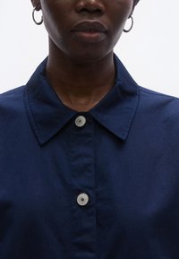 Sötétkék galléros ing gombos záródással, sima szövetből készült. Ezüst gombdíszítéssel és strukturált gallérral rendelkezik.