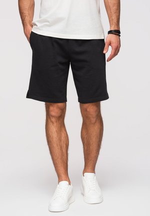 Uomo che indossa pantaloncini neri fino al ginocchio, scarpe da ginnastica bianche e una camicia bianca, con le mani rilassate ai lati su uno sfondo chiaro e uniforme.