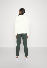Persona con capelli scuri raccolti, indossa una maglietta bianca a maniche lunghe, pantaloni verdi scuri e sneaker bianchi, di fronte a un semplice muro grigio chiaro.