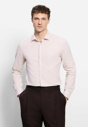 Jeune homme portant une chemise rose clair à manches longues boutonnée et un pantalon marron foncé, debout les mains dans les poches sur fond blanc.