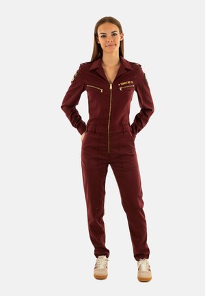 Una donna sta in piedi con le mani nelle tasche, indossando una tuta zip-up bordeaux con toppe e sneakers beige, contro uno sfondo semplice.