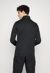 Cămașă polo neagră cu mâneci lungi și guler, fabricată dintr-un material neted. Prezintă un design minimalist și o formă ajustată, văzută din spate.