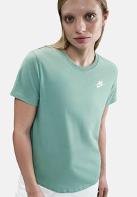 Femme portant un t-shirt Nike vert menthe avec le logo blanc sur la poitrine, se tenant devant un fond clair.