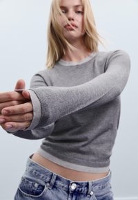 Grauer Strickpullover mit rundem Ausschnitt, gerippten Bündchen und einem kontrastierenden hellgrauen Saum. Weiche Textur, die in einer lässigen Pose zur Geltung kommt.